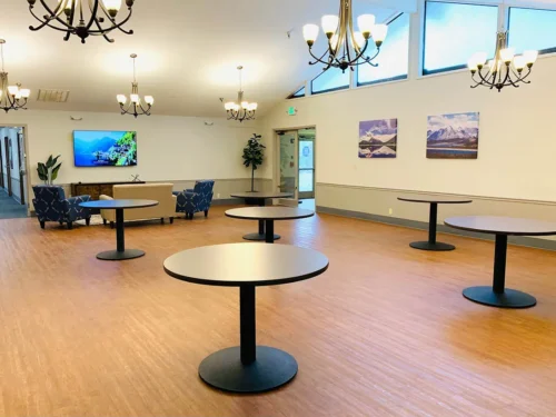 Common area with wooden flooring, round tables, chandeliers, wall art, and a seating area with TV