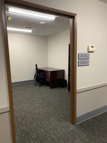 Office room with carpet flooring, desk, chair, and sign reading “Director of Health Services” and “Resident Care Coordinator
